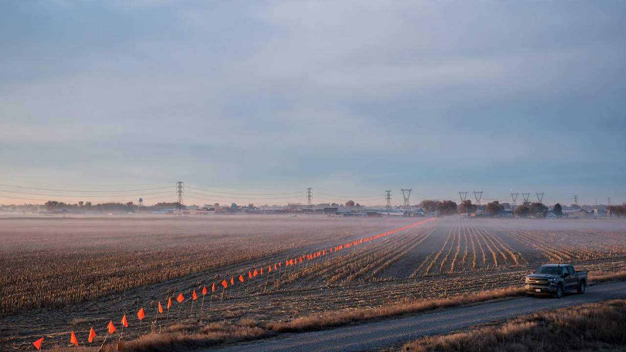Orange survey flags cross a harvested field toward distant power lines at dawn, a lone pickup parked on the shoulder.