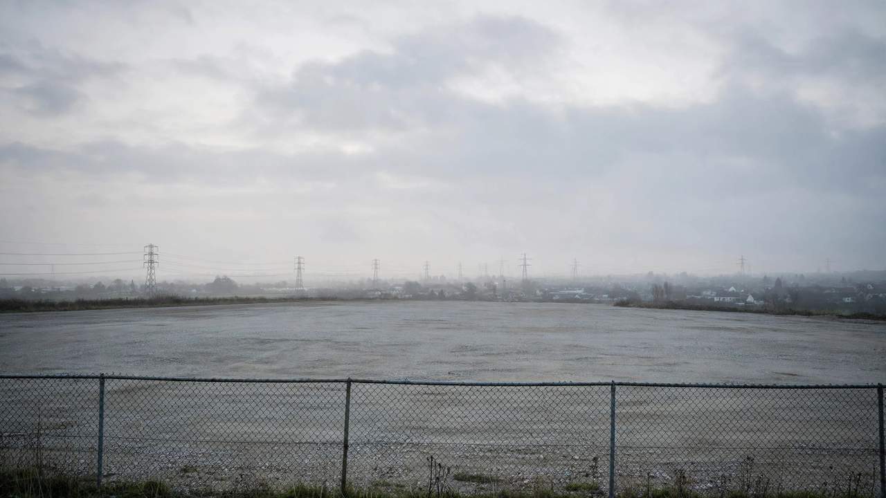 Cleared gravel lot behind a chain-link fence, with distant power pylons fading into morning haze.